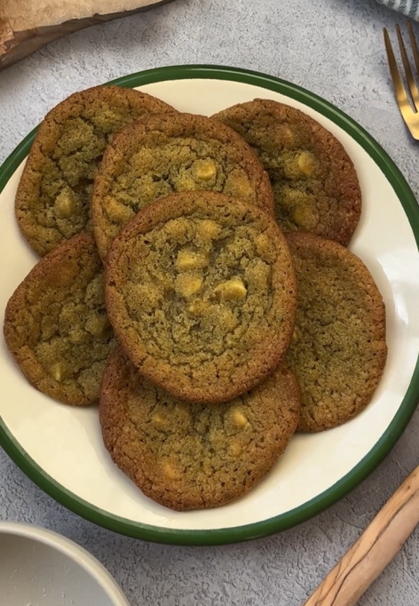 Ein Teller mit Matcha-Cookies mit weißen Schokoladenstückchen steht auf einem grauen Tisch neben einer goldenen Gabel und einem Messer, einer gestreiften Serviette und einem Holzbrett.