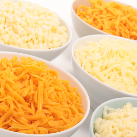 Fünf weiße Schalen, gefüllt mit verschiedenen Sorten geraspelten und gewürfelten Käses, darunter gelber Cheddar und weißer Käse, sind auf einer weißen Fläche angeordnet.