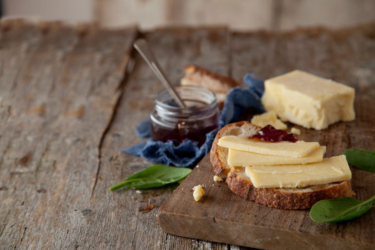 Eine Scheibe rustikales Brot mit altem Cheddar-Käse und Marmelade liegt auf einem Holzbrett neben Spinatblättern; im Hintergrund stehen ein Glas Marmelade mit einem Löffel und ein Block Käse.