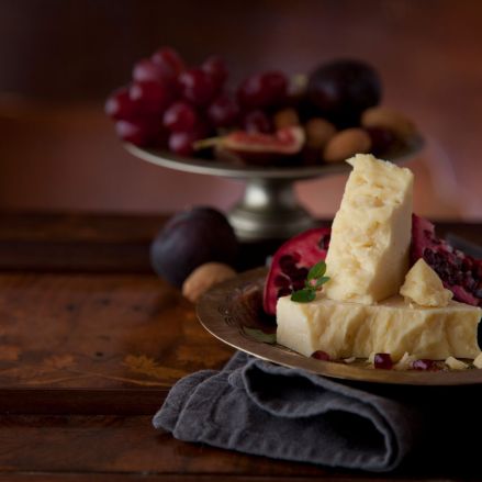 Ein Teller mit Cheddar-Käse, Granatapfel und Trauben steht auf einem Holztisch. Im Hintergrund stehen auf einem weiteren Teller Trauben, Feigen und Mandeln, die ein rustikales, elegantes Arrangement ergeben.