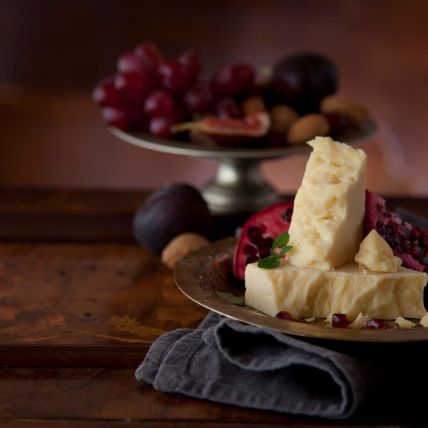 Ein Teller mit Cheddar-Käse, Granatapfel und Trauben steht auf einem Holztisch. Im Hintergrund stehen auf einem weiteren Teller Trauben, Feigen und Mandeln, die ein rustikales, elegantes Arrangement ergeben.