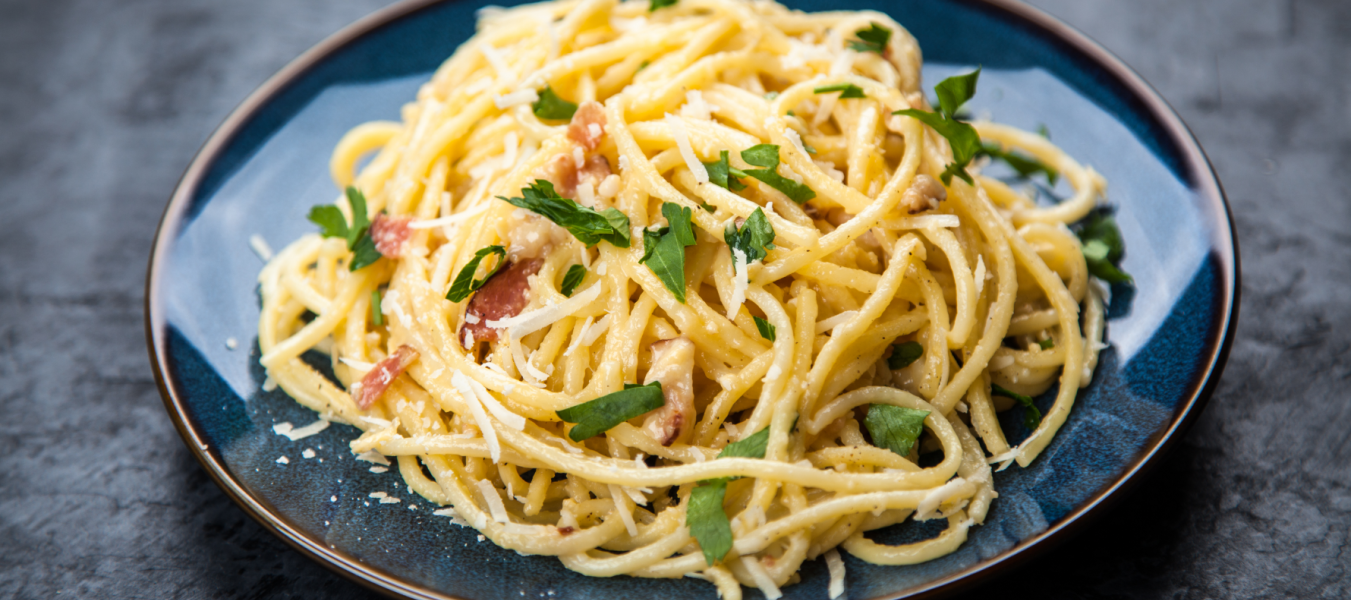 Ein Teller Spaghetti Carbonara mit gehackter Petersilie und geriebenem Käse steht auf einem blauen Teller vor einem dunklen, strukturierten Hintergrund, daneben eine Scheibe herzhaftes French Toast für eine schmackhafte Abwechslung.