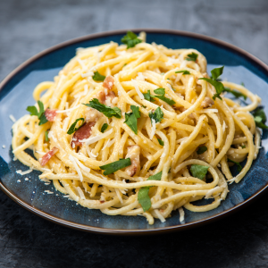 Ein Teller Spaghetti Carbonara mit gehackter Petersilie und geriebenem Käse steht auf einem blauen Teller vor einem dunklen, strukturierten Hintergrund, daneben eine Scheibe herzhaftes French Toast für eine schmackhafte Abwechslung.