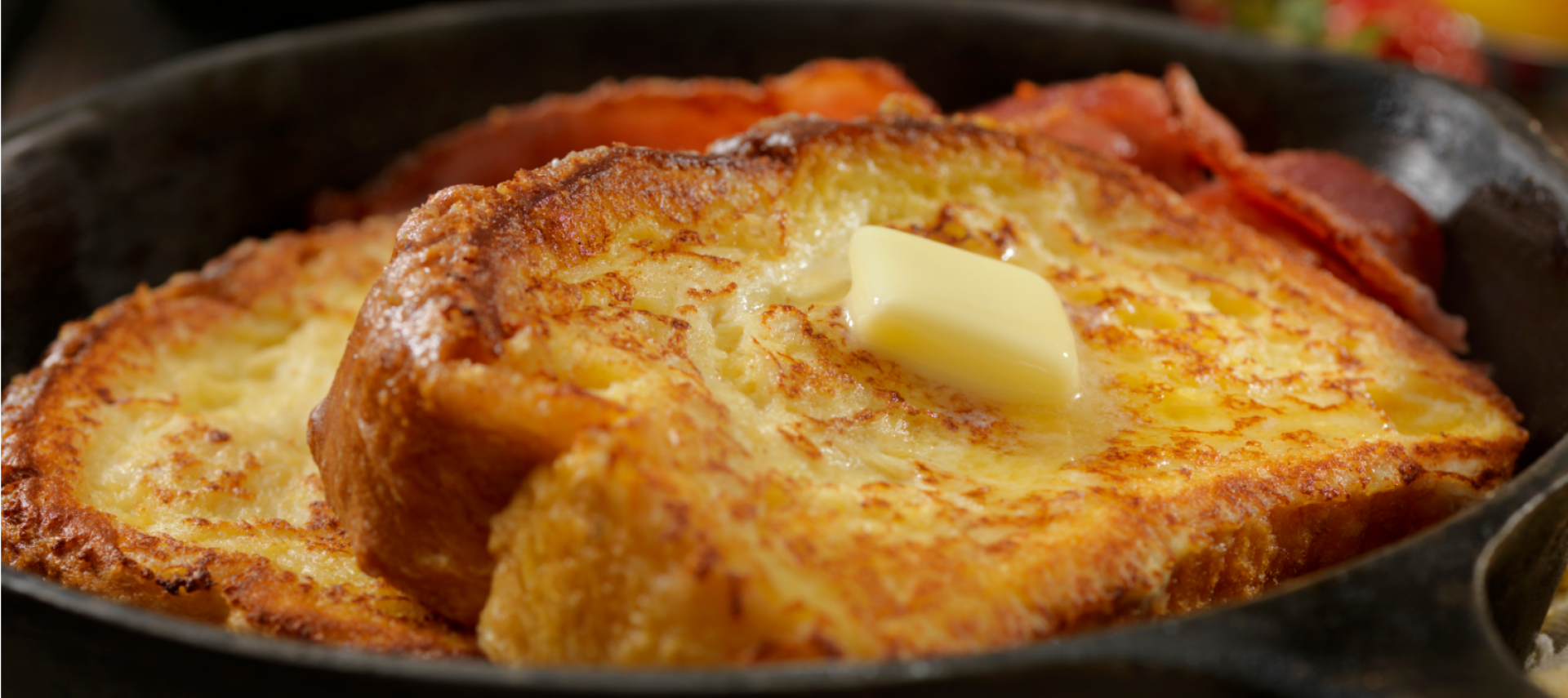 Zwei Scheiben French Toast mit Honig-Butter obenauf, serviert in einer gusseisernen Pfanne mit Streifen von knusprigem Speck. Im Hintergrund gibt es Erdbeeren und ein Glas Orangensaft für ein perfektes herzhaftes Frühstück.