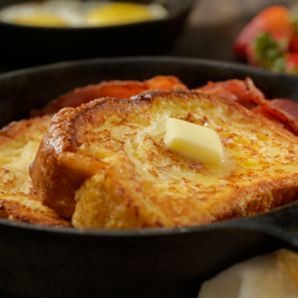 Zwei Scheiben French Toast mit Honig-Butter obenauf, serviert in einer gusseisernen Pfanne mit Streifen von knusprigem Speck. Im Hintergrund gibt es Erdbeeren und ein Glas Orangensaft für ein perfektes herzhaftes Frühstück.