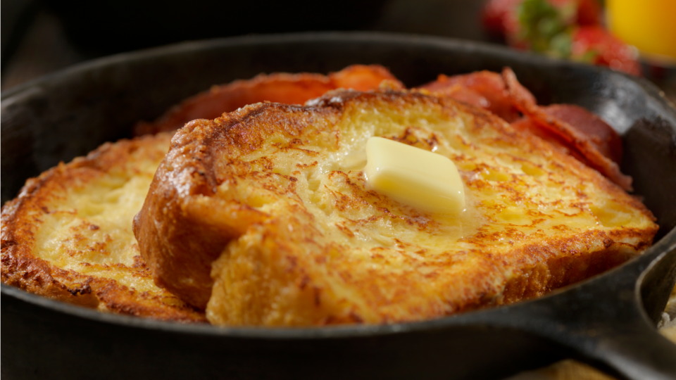 Zwei Scheiben French Toast mit Honig-Butter obenauf, serviert in einer gusseisernen Pfanne mit Streifen von knusprigem Speck. Im Hintergrund gibt es Erdbeeren und ein Glas Orangensaft für ein perfektes herzhaftes Frühstück.