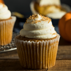 Ein Kürbis-Cupcake mit weißem Zuckerguss und braunem Zucker steht auf einem Holztisch, der eine gemütliche French-Toast-Atmosphäre ausstrahlt. Ein zweiter Cupcake und ein kleiner Kürbis sind unscharf im Hintergrund zu sehen.