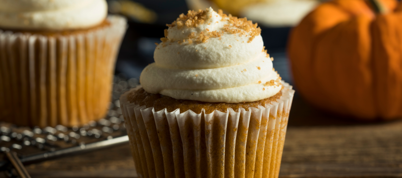 Ein Kürbis-Cupcake mit weißem Zuckerguss und braunem Zucker steht auf einem Holztisch, der eine gemütliche French-Toast-Atmosphäre ausstrahlt. Ein zweiter Cupcake und ein kleiner Kürbis sind unscharf im Hintergrund zu sehen.