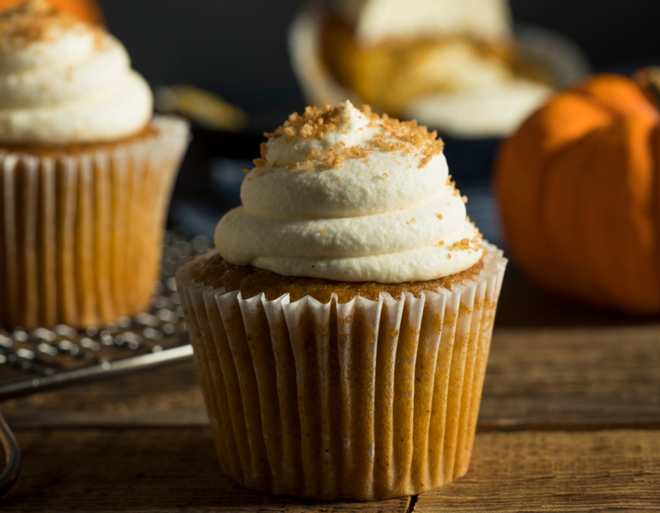 Ein Kürbis-Cupcake mit weißem Zuckerguss und braunem Zucker steht auf einem Holztisch, der eine gemütliche French-Toast-Atmosphäre ausstrahlt. Ein zweiter Cupcake und ein kleiner Kürbis sind unscharf im Hintergrund zu sehen.