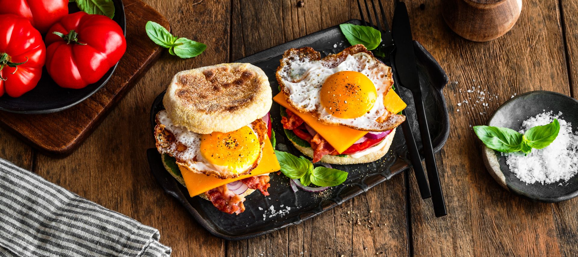 Frühstückssandwich mit Spiegeleiern, Cheddarkäse, Speck und Salat auf englischen Muffins werden auf einem schwarzen Tablett mit Basilikumblättern serviert