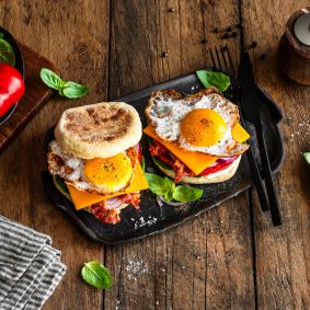 Frühstückssandwich mit Spiegeleiern, Cheddarkäse, Speck und Salat auf englischen Muffins werden auf einem schwarzen Tablett mit Basilikumblättern serviert