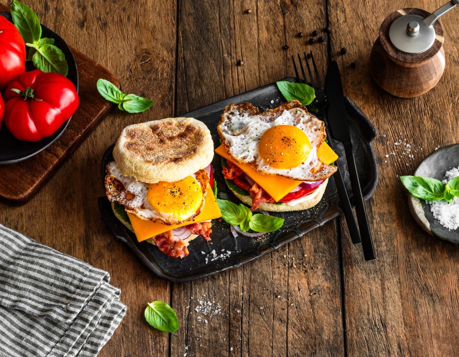 Frühstückssandwich mit Spiegeleiern, Cheddarkäse, Speck und Salat auf englischen Muffins werden auf einem schwarzen Tablett mit Basilikumblättern serviert