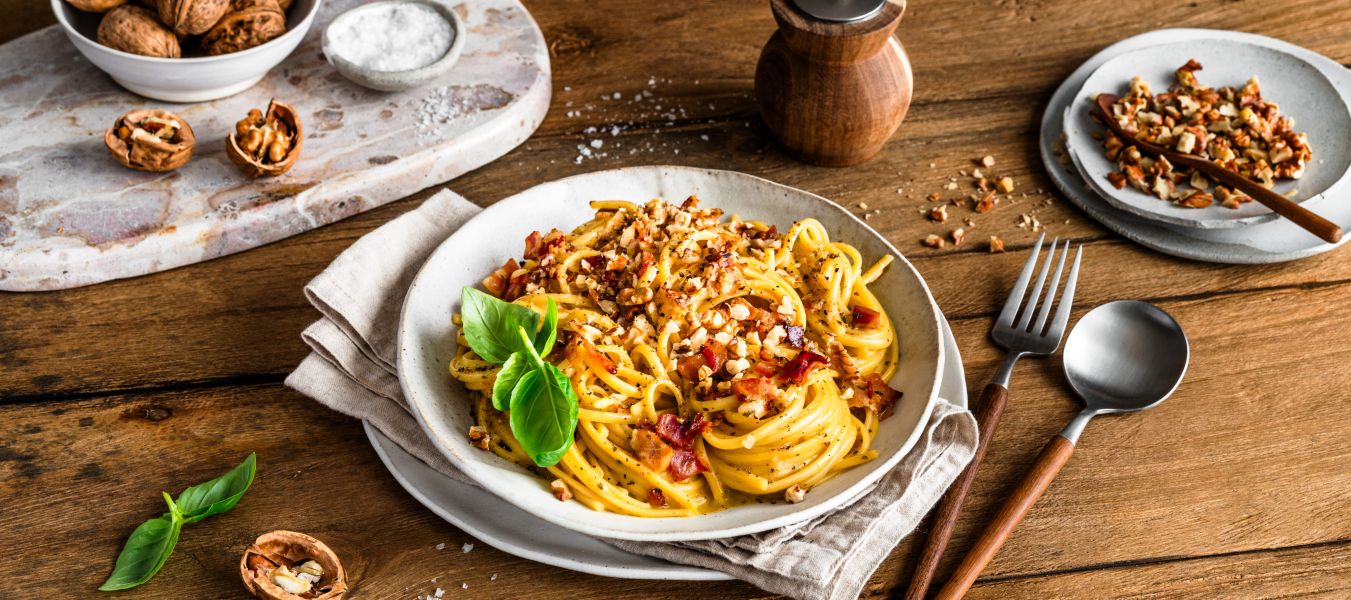 Ein Teller mit Spaghetti Carbonara, garniert mit gehackten Walnüssen und frischen Basilikumblättern, steht auf einem rustikalen Holztisch. Daneben liegen eine Gabel und ein Löffel mit Holzgriffen auf einer Serviette.