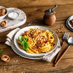 Ein Teller mit Spaghetti Carbonara, garniert mit gehackten Walnüssen und frischen Basilikumblättern, steht auf einem rustikalen Holztisch. Daneben liegen eine Gabel und ein Löffel mit Holzgriffen auf einer Serviette.