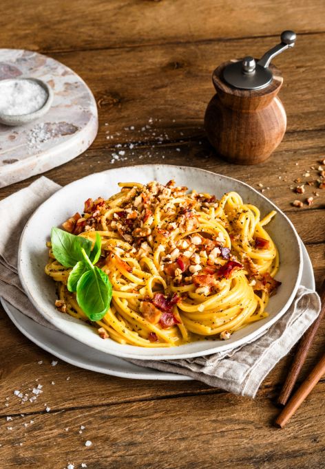 Ein Teller mit Spaghetti Carbonara, garniert mit gehackten Walnüssen und frischen Basilikumblättern, steht auf einem rustikalen Holztisch. Daneben liegen eine Gabel und ein Löffel mit Holzgriffen auf einer Serviette.