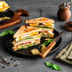 Ein Club-Sandwich, diagonal halbiert und mit Metallspießen fixiert, liegt auf einem schwarzen Teller. Die Sandwich-Hälften bestehen aus getoastetem Brot, Salat, Tomate, Käse, Schinken oder Putenaufschnitt und Ei. Um den Teller herum sind Kartoffelchips und frische Basilikumblätter verteilt.