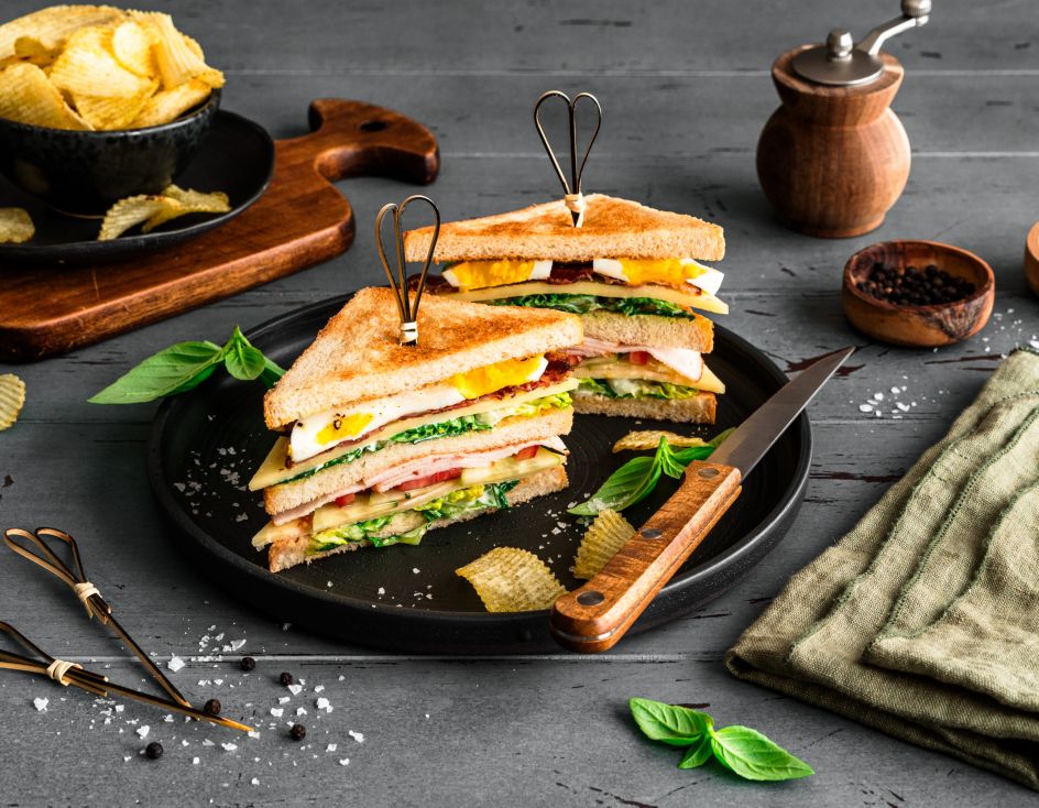 Ein Club-Sandwich, diagonal halbiert und mit Metallspießen fixiert, liegt auf einem schwarzen Teller. Die Sandwich-Hälften bestehen aus getoastetem Brot, Salat, Tomate, Käse, Schinken oder Putenaufschnitt und Ei. Um den Teller herum sind Kartoffelchips und frische Basilikumblätter verteilt.