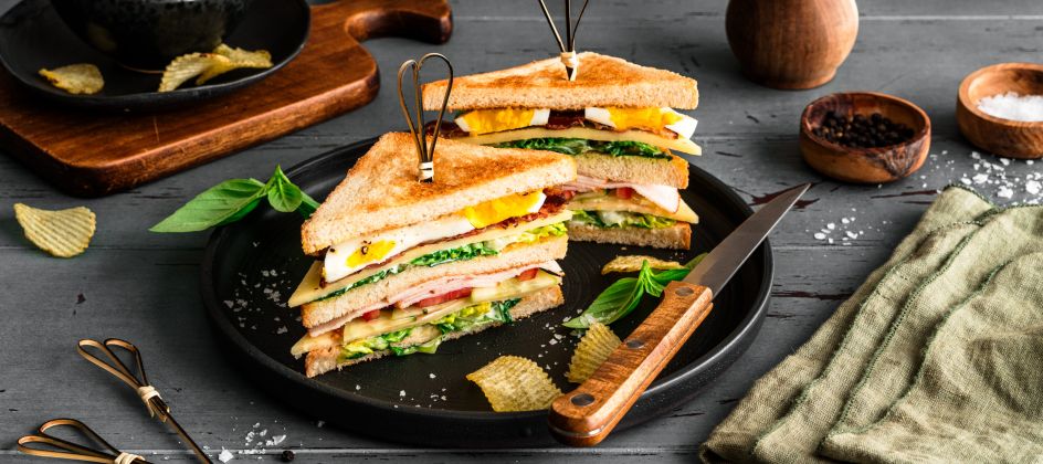 Ein Club-Sandwich, diagonal halbiert und mit Metallspießen fixiert, liegt auf einem schwarzen Teller. Die Sandwich-Hälften bestehen aus getoastetem Brot, Salat, Tomate, Käse, Schinken oder Putenaufschnitt und Ei. Um den Teller herum sind Kartoffelchips und frische Basilikumblätter verteilt.