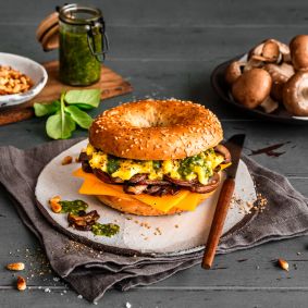Ein Frühstücksbagel mit Rührei, Cheddarkäse, Champignons und Pesto liegt auf einem Teller. Rohe Champignons, grünes Pesto, Pinienkerne und frische Basilikumblätter umgeben das Sandwich auf einem rustikalen Tisch.