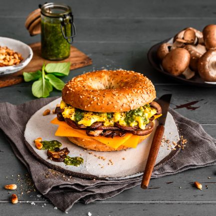 Ein Frühstücksbagel mit Rührei, Cheddarkäse, Champignons und Pesto liegt auf einem Teller. Rohe Champignons, grünes Pesto, Pinienkerne und frische Basilikumblätter umgeben das Sandwich auf einem rustikalen Tisch.