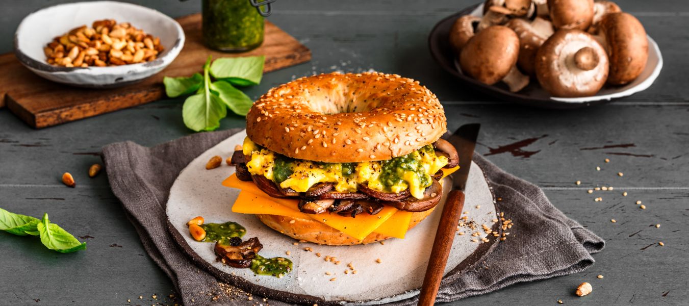 Ein Frühstücksbagel mit Rührei, Cheddarkäse, Champignons und Pesto liegt auf einem Teller. Rohe Champignons, grünes Pesto, Pinienkerne und frische Basilikumblätter umgeben das Sandwich auf einem rustikalen Tisch.