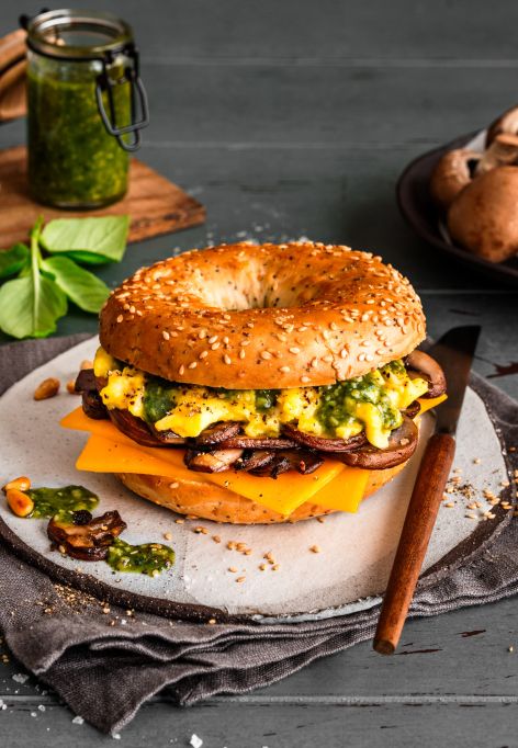 Ein Frühstücksbagel mit Rührei, Cheddarkäse, Champignons und Pesto liegt auf einem Teller. Rohe Champignons, grünes Pesto, Pinienkerne und frische Basilikumblätter umgeben das Sandwich auf einem rustikalen Tisch.