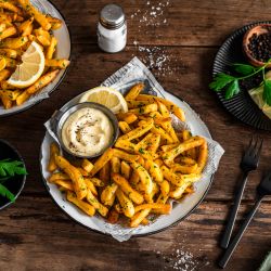 Auf einem Holztisch stehen Teller mit gewürzten Pommes, garniert mit gehackter Petersilie und Zitronenspalten. Auf einem Teller befindet sich zusätzlich ein kleines Metallschälchen mit Aioli.