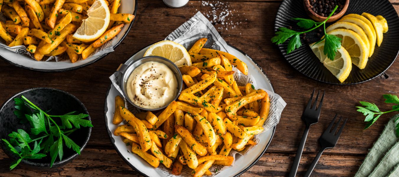 Auf einem Holztisch stehen Teller mit gewürzten Pommes, garniert mit gehackter Petersilie und Zitronenspalten. Auf einem Teller befindet sich zusätzlich ein kleines Metallschälchen mit Aioli.