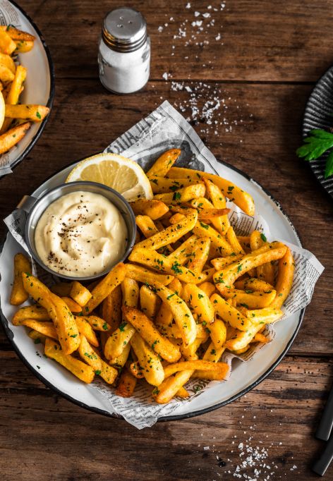 Auf einem Holztisch stehen Teller mit gewürzten Pommes, garniert mit gehackter Petersilie und Zitronenspalten. Auf einem Teller befindet sich zusätzlich ein kleines Metallschälchen mit Aioli.