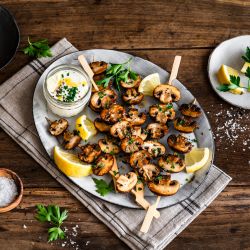 Ein Teller mit gegrillten Pilzspießen, garniert mit gehackter Petersilie und Zitronenspalten. Auf dem Teller steht außerdem ein kleines Glas mit einem cremigen Dip, der mit Kräutern bestreut ist.