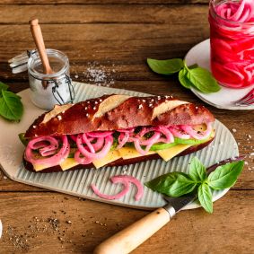 Ein Sandwich mit Laugenbrötchen, Käse, Salat und eingelegten roten Zwiebeln liegt auf einem Holzbrett. Daneben stehen Gewürze, Basilikum und ein Glas mit eingelegten Zwiebeln.