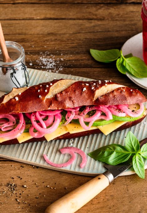 Ein Sandwich mit Laugenbrötchen, Käse, Salat und eingelegten roten Zwiebeln liegt auf einem Holzbrett. Daneben stehen Gewürze, Basilikum und ein Glas mit eingelegten Zwiebeln.