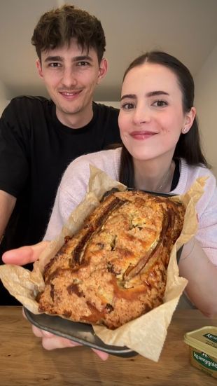 Eine lächelnde Frau hält einen Laib selbstgebackenes Bananenbrot auf Pergamentpapier, während ein Mann neben ihr steht.
