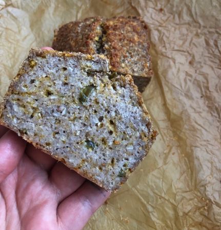 Eine Hand hält eine dicke, leckere Scheibe rustikales Brot mit sichtbaren Samen und Körnern. Im Hintergrund liegt weiteres Brot auf zerknittertem Pergamentpapier.
