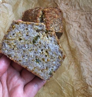 Eine Hand hält eine dicke, leckere Scheibe rustikales Brot mit sichtbaren Samen und Körnern. Im Hintergrund liegt weiteres Brot auf zerknittertem Pergamentpapier.