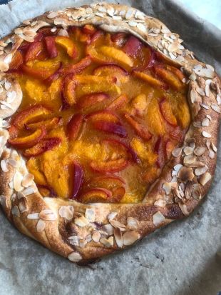 Eine rustikale, köstliche Fruchtgalette mit einer goldgelben, gefalteten Kruste, die mit gehobelten Mandeln belegt und mit saftigem gelbem und rotem Steinobst gefüllt ist, gebacken auf Pergamentpapier.