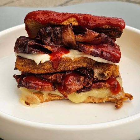 Eine Nahaufnahme eines Sandwiches mit knusprigem Speck, geschmolzenem Käse und Brotscheiben, belegt mit einem Spritzer Ketchup, serviert auf einem weißen Teller von nik_prins.