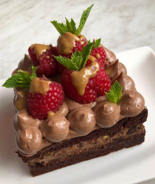 Ein quadratisches Stück Schokoladenkuchen mit Mousse au Chocolat, frischen Himbeeren, Minzblättern und einer goldenen Sauce, serviert auf einem weißen Teller.