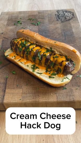 Eine gegrillte Wurst, belegt mit geschmolzenem Käse und gehackten Kräutern, liegt in einem Brötchen auf einem Holzschneidebrett. Der Text am unteren Rand lautet Cream Cheese Hack Dog von bbqdad85.