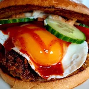 Nahaufnahme eines frisch gegrillten Burgers mit Spiegelei, flüssigem Eigelb, Ketchup oder Soße, Gurkenscheiben, Salat und einem Burger-Patty in einem getoasteten Brötchen.