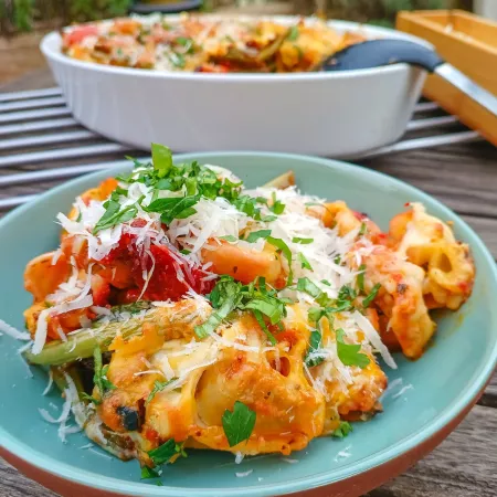 Ein weißer Teller mit einer Portion gebackener Pasta, bedeckt mit geschmolzenem geriebenem Käse und frischen grünen Kräutern. Im Hintergrund steht eine rechteckige Auflaufform mit weiterer Pasta derselben Art auf einem Holztisch.