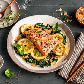 Gebratenes Lachsfilet auf sautiertem Spinat mit Zitronenscheiben, garniert mit Kräutern und gerösteten Mandelblättchen. Serviert auf einem gedeckten Tisch mit Salz, Pfeffer, Mandeln und frischem Basilikum, daneben ein Besteckset auf einer gestreiften Stoffserviette.