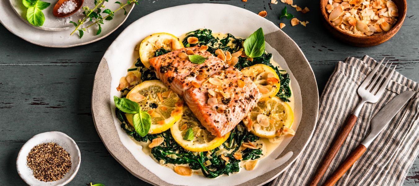 Gebratenes Lachsfilet auf sautiertem Spinat mit Zitronenscheiben, garniert mit Kräutern und gerösteten Mandelblättchen. Serviert auf einem gedeckten Tisch mit Salz, Pfeffer, Mandeln und frischem Basilikum, daneben ein Besteckset auf einer gestreiften Stoffserviette.