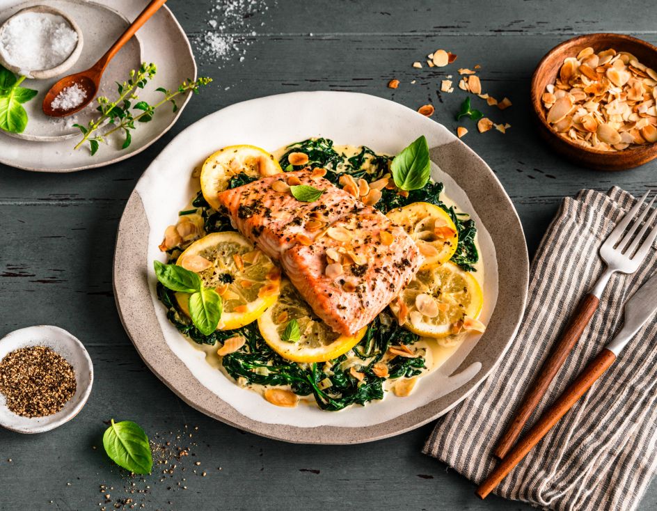 Gebratenes Lachsfilet auf sautiertem Spinat mit Zitronenscheiben, garniert mit Kräutern und gerösteten Mandelblättchen. Serviert auf einem gedeckten Tisch mit Salz, Pfeffer, Mandeln und frischem Basilikum, daneben ein Besteckset auf einer gestreiften Stoffserviette.