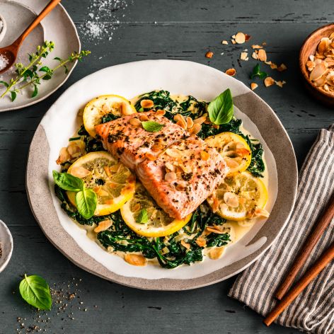 Gebratenes Lachsfilet auf sautiertem Spinat mit Zitronenscheiben, garniert mit Kräutern und gerösteten Mandelblättchen. Serviert auf einem gedeckten Tisch mit Salz, Pfeffer, Mandeln und frischem Basilikum, daneben ein Besteckset auf einer gestreiften Stoffserviette.
