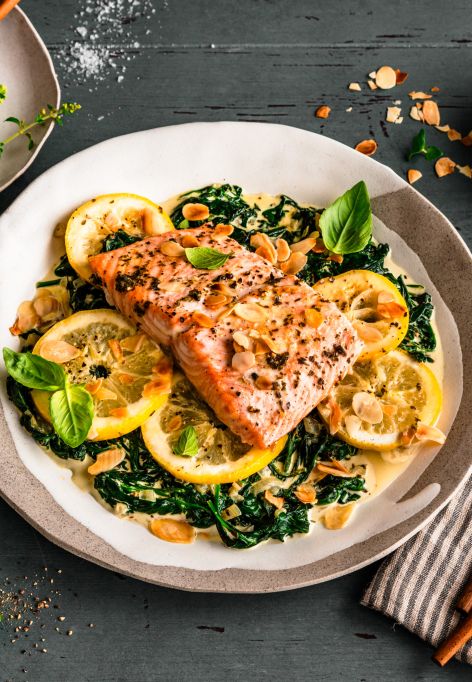 Gebratenes Lachsfilet auf sautiertem Spinat mit Zitronenscheiben, garniert mit Kräutern und gerösteten Mandelblättchen. Serviert auf einem gedeckten Tisch mit Salz, Pfeffer, Mandeln und frischem Basilikum, daneben ein Besteckset auf einer gestreiften Stoffserviette.