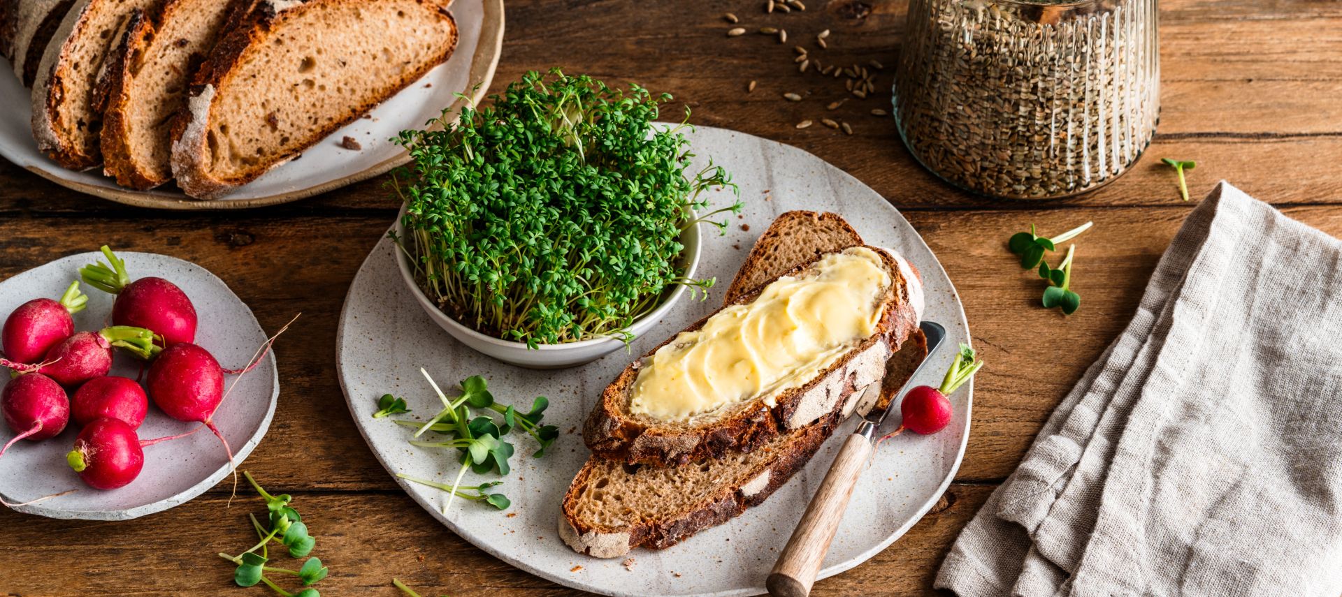 Auf einem rustikalen Holztisch liegen mehrere Scheiben knuspriges Brot, frische Radieschen, eine mit Butter bestrichene Brotscheibe, eine kleine Schale mit grünen Sprossen, ein Glas mit Körnern sowie eine gefaltete beigefarbene Stoffserviette.