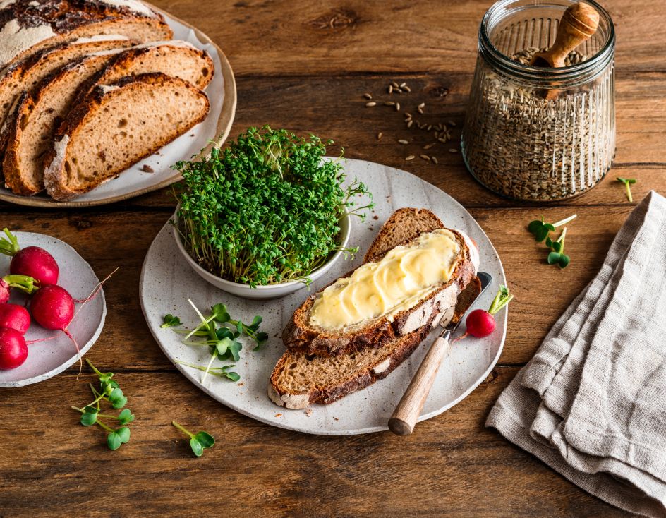 Auf einem rustikalen Holztisch liegen mehrere Scheiben knuspriges Brot, frische Radieschen, eine mit Butter bestrichene Brotscheibe, eine kleine Schale mit grünen Sprossen, ein Glas mit Körnern sowie eine gefaltete beigefarbene Stoffserviette.