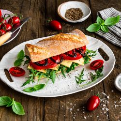 Frisches Ciabatta auf einem weißen Teller, belegt mit Rucola, Käsescheiben und Tomaten. Auf dem Holztisch rundherum liegen Kirschtomaten, Basilikumblätter, Salz und Pfeffer in kleinen Schalen sowie eine gestreifte Stoffserviette.