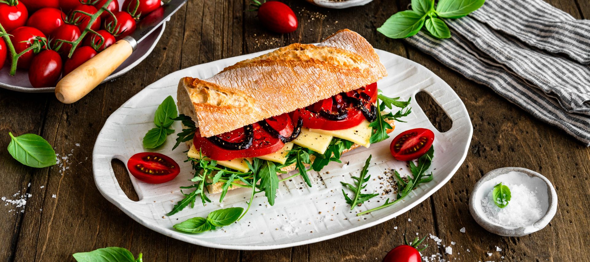 Frisches Ciabatta auf einem weißen Teller, belegt mit Rucola, Käsescheiben und Tomaten. Auf dem Holztisch rundherum liegen Kirschtomaten, Basilikumblätter, Salz und Pfeffer in kleinen Schalen sowie eine gestreifte Stoffserviette.